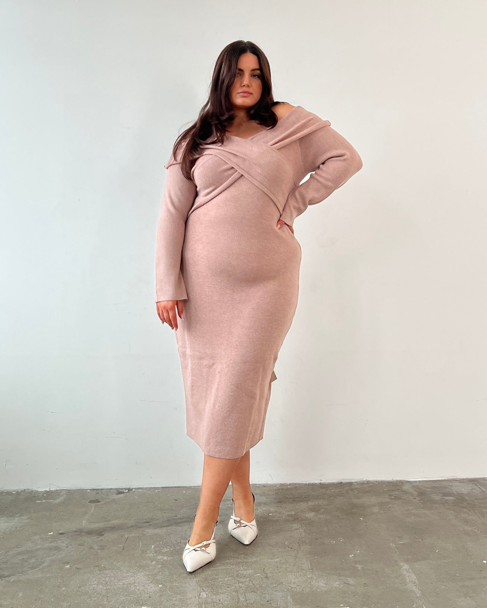 Woman wearing a pink dress and wide fit white heels standing against a white wall.