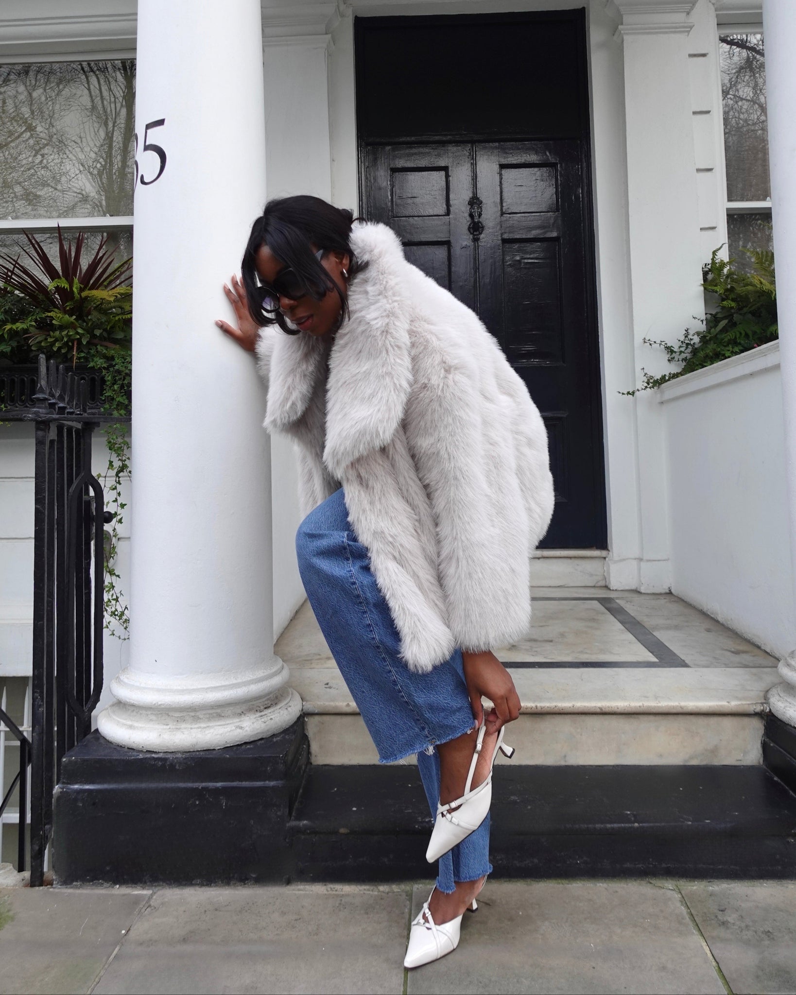 Woman wearing wide fit white heels in a white fur coat and blue jeans standing on a porch.
