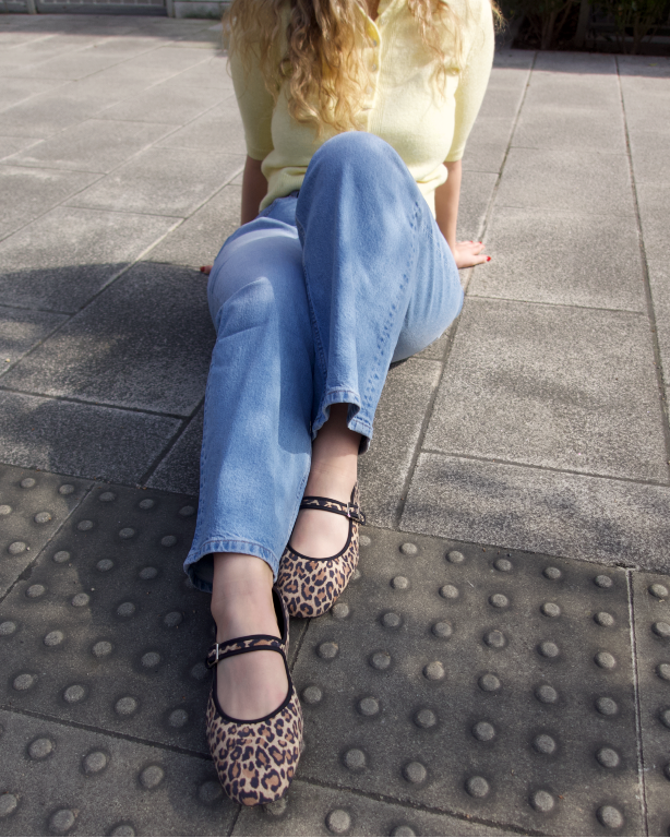 Person sitting on a sidewalk wearing wide fit leopard print shoes, blue jeans, and a yellow top.