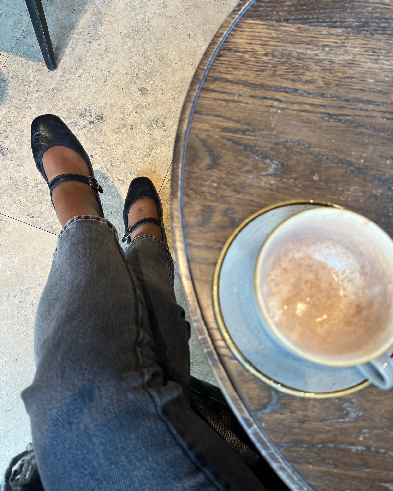 Person wearing wide fit black flats and gray pants standing next to a wooden table with a coffee cup on it.